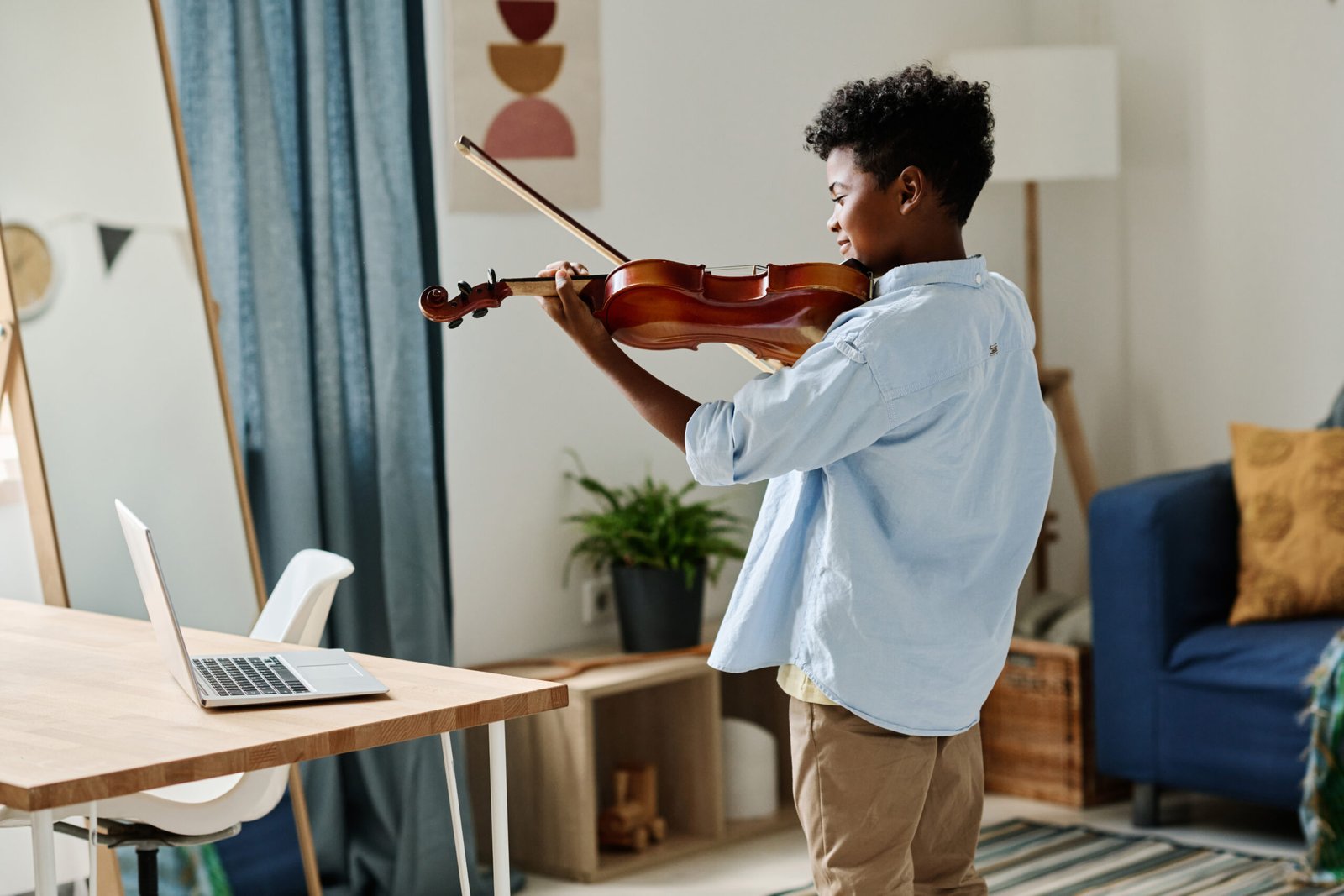 Rear view of African boy standing in his room and practicing to play violin during online lesson on laptop